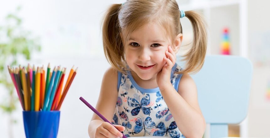 Ein kleines Mädchen sitzt lächelnd an einem Tisch und malt mit Buntstiften